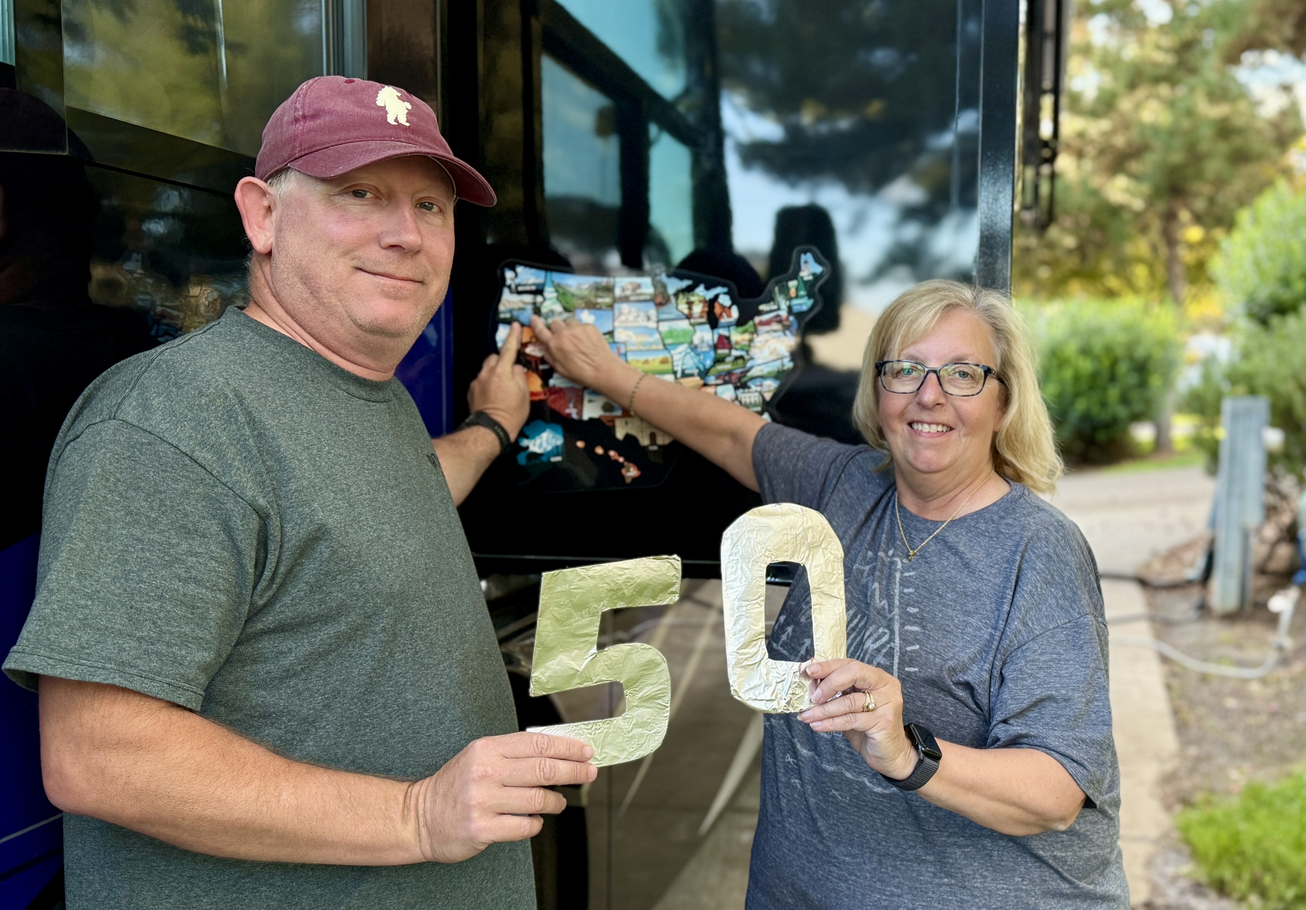 Chad and Donna outside their motorhome — Donna points to the RV's fifty-state sticker map while they hold up a gold number 50.
