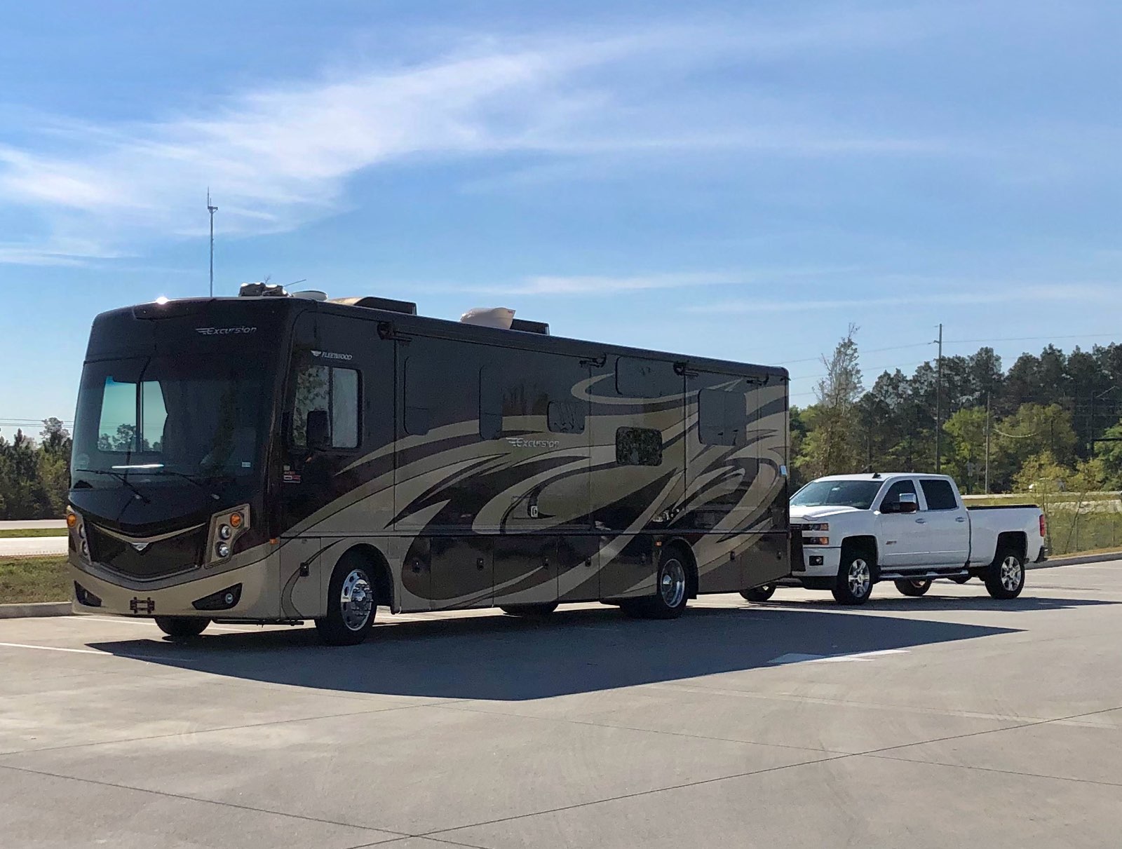 2016 Fleetwood Excursion 35E Class A motorhome with a white Chevy Silverado toad parked at a rest area under a blue sky