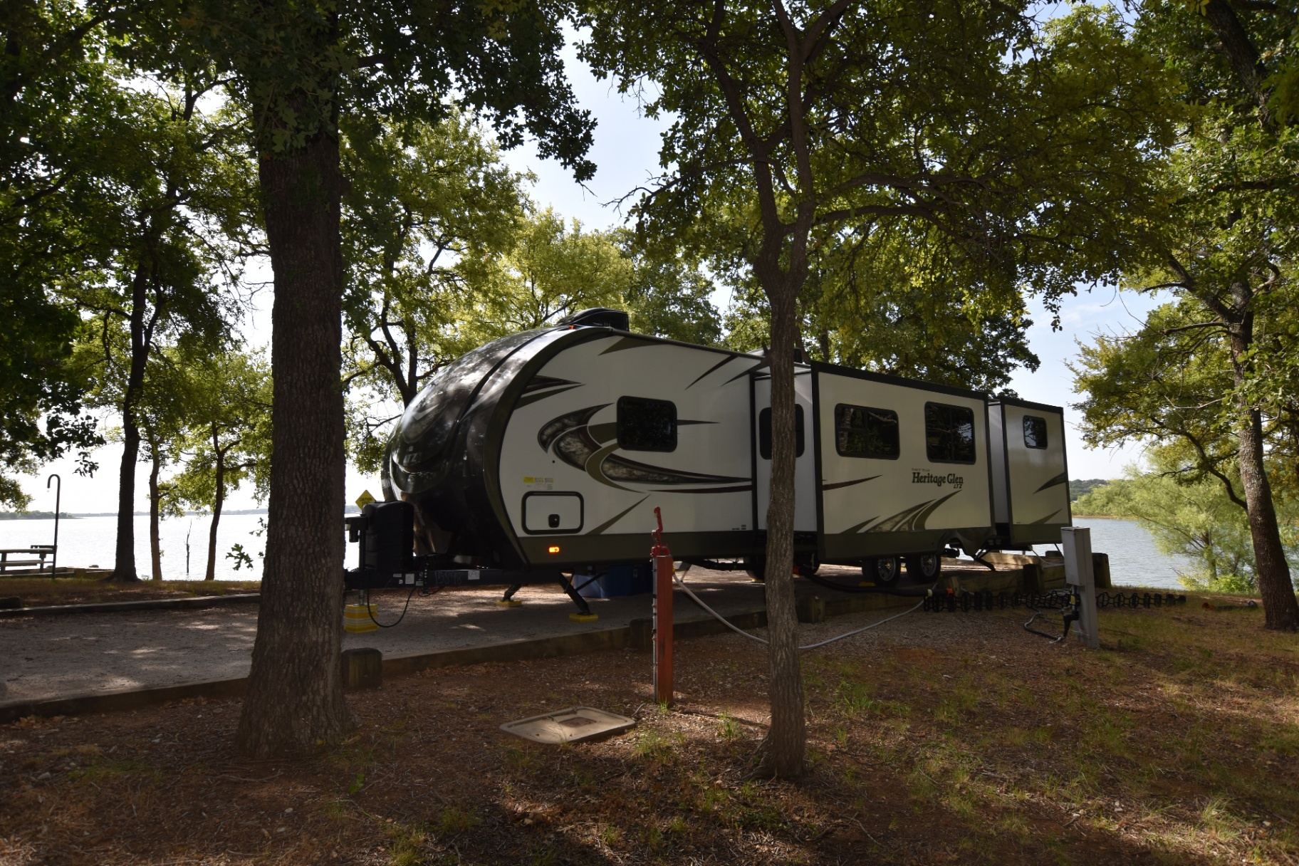 2018 Forest River Heritage Glen LTZ 312QBUD parked at a lakeside campsite framed by hardwood trees