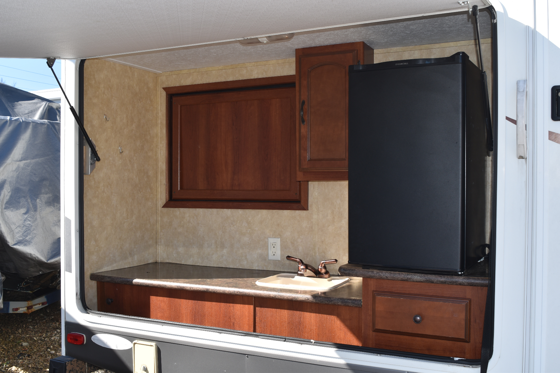 Heritage Glen exterior outdoor kitchen bay open showing the stainless sink, countertop, cabinet, and black mini-fridge