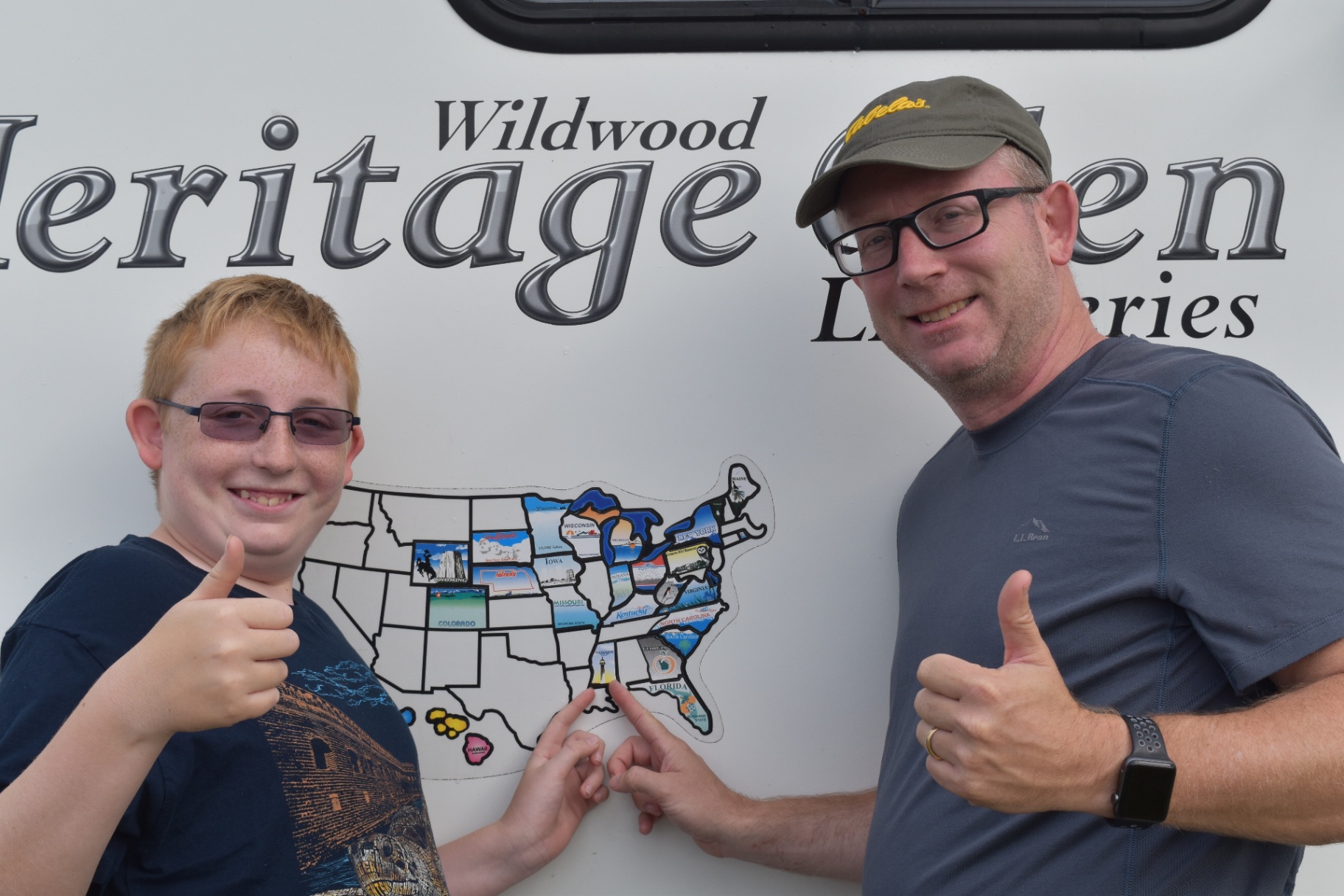 Father and son giving a thumbs-up in front of the USA state-by-state travel sticker map mounted on the Heritage Glen